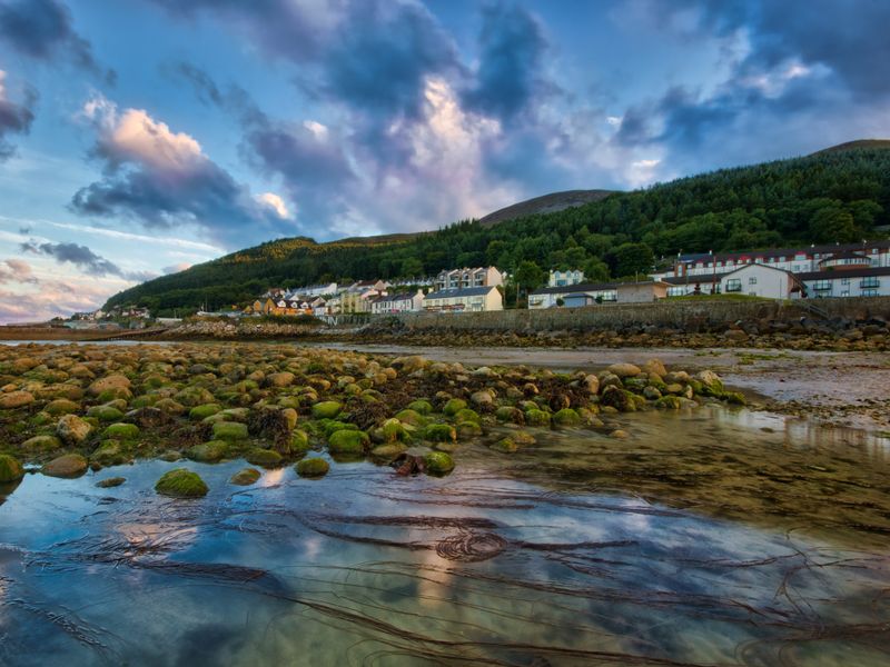 Most likely, you'll start your Slieve Donard hike from Newcastle. This image shows seaside houses against the backdrop of a forest and mountain tops.